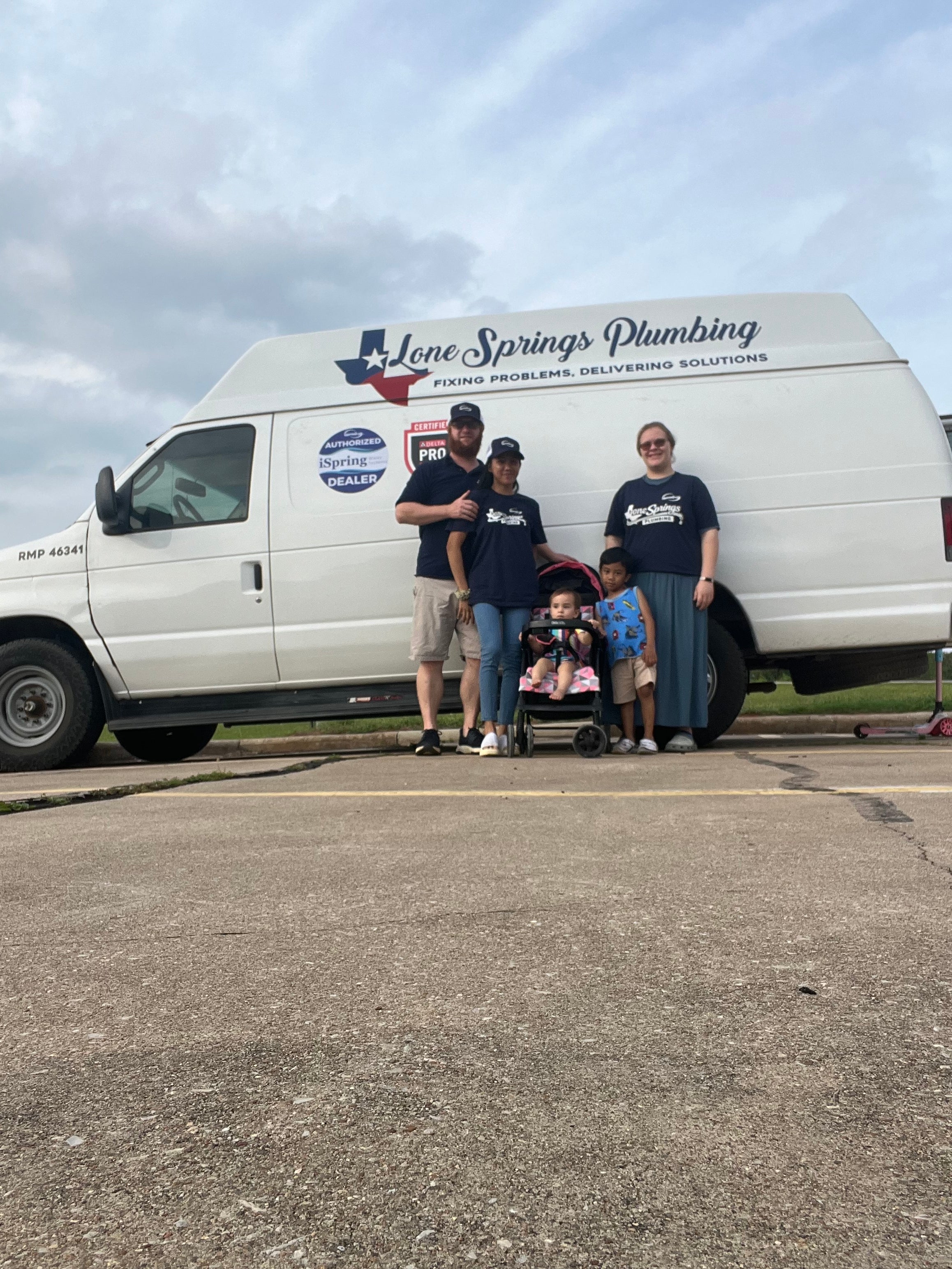 A group of people posing in front of a white service van with 'Lane Springs Plumbing' printed on the side, parked outdoors on a paved surface under a cloudy sky.